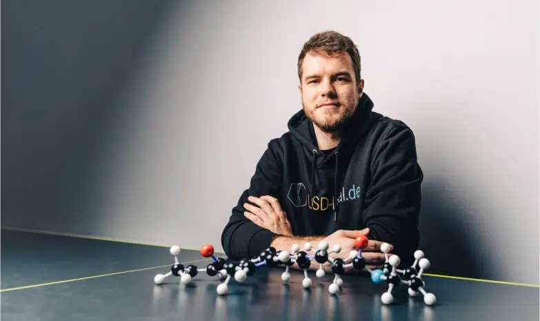 A man is posing in front of a table full of molecule models and wearing a black hoodie with the lsd legal label on it.